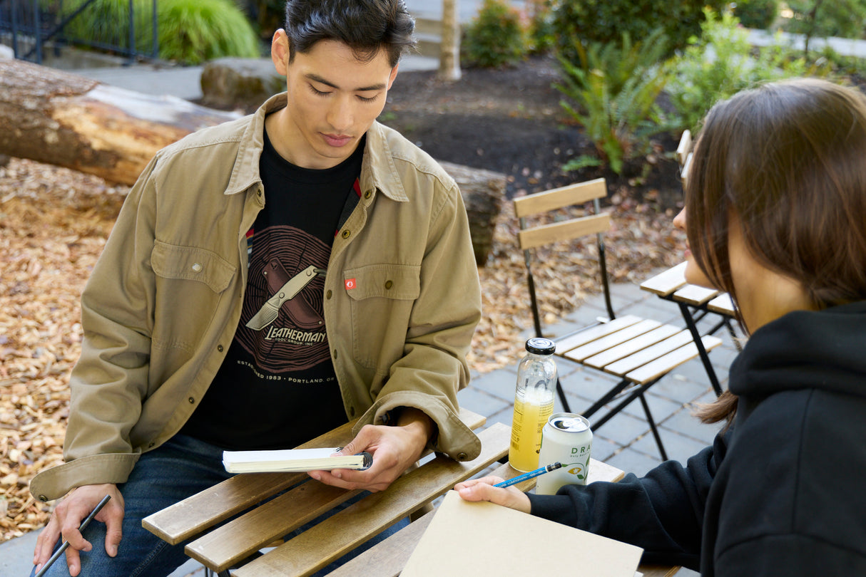 Translation missing: en.A man sitting at a table wearing a black Leatherman Blade & Grain tee with a woman wearing a black hoodie, both equipped with a notebook and pencil