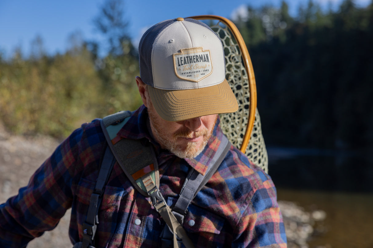 Translation missing: en.A person wearing a tan and brown Leatherman PNW Trucker Hat standing near a river with a backpack and fishing net