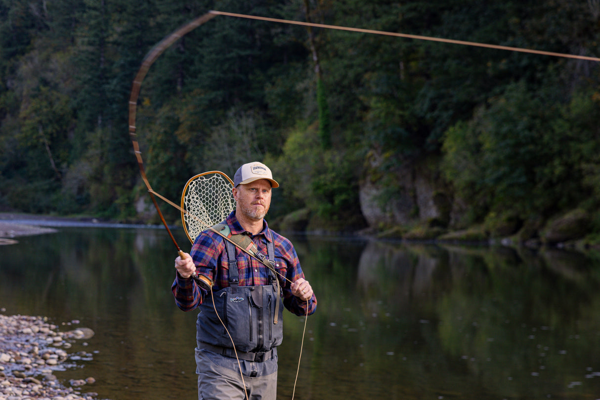 Translation missing: en.A person wearing a beige Leatherman PNW Trucker hat fly-fishing in a river surrounded by trees