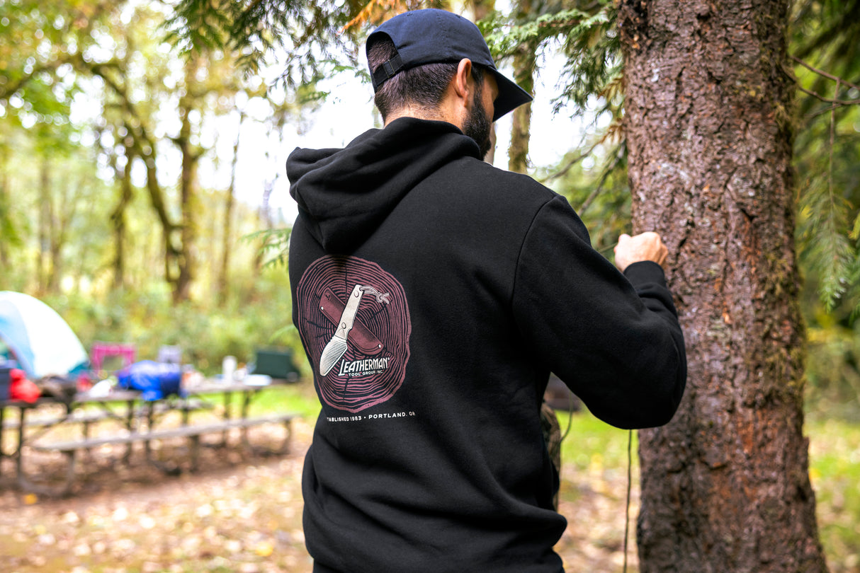 Translation missing: en.A person wearing a black Leatherman Blade &amp; Grain hoodie standing next to a tree in a forest campsite. The hoodie’s back graphic shows a pocketknife illustration with “Leatherman Tool Group” text