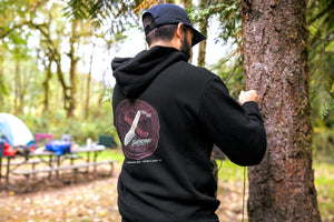 Translation missing: en.A person wearing a black Leatherman Blade &amp; Grain hoodie standing next to a tree in a forest campsite. The hoodie’s back graphic shows a pocketknife illustration with “Leatherman Tool Group” text
