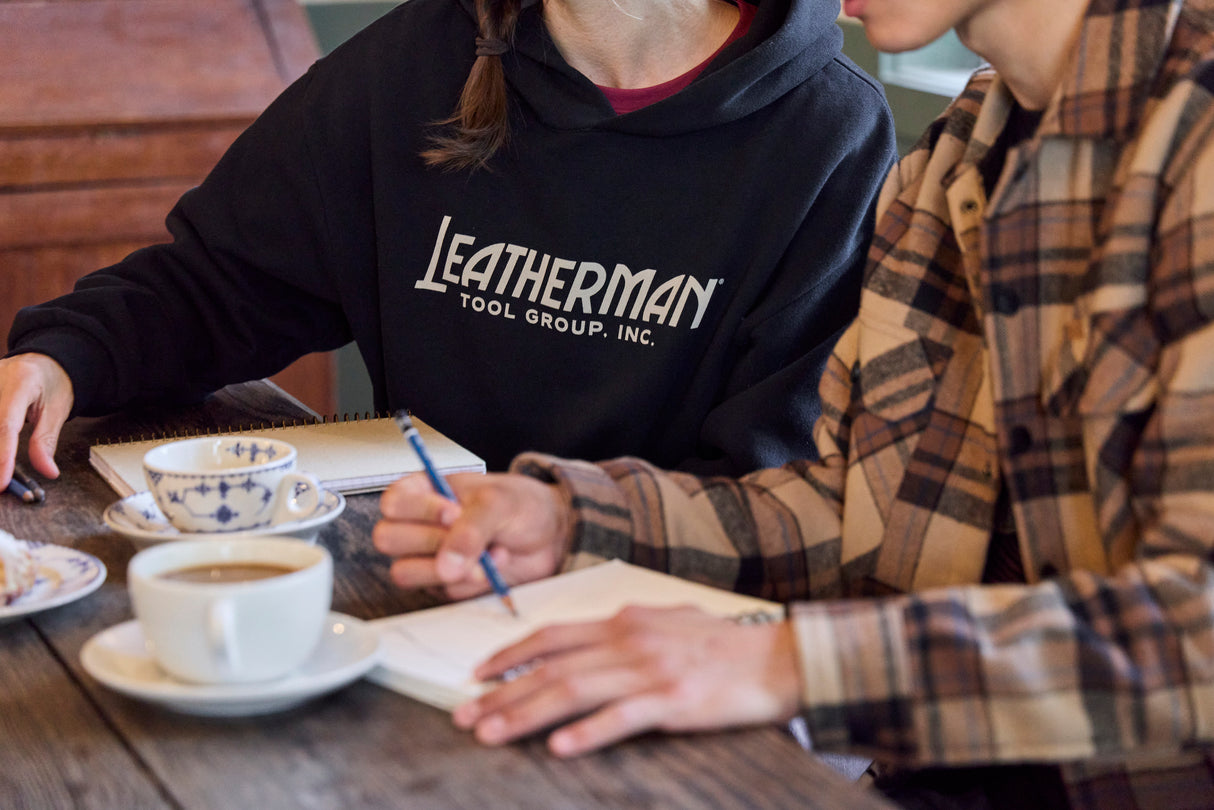 Translation missing: en.Two people sitting at a table with coffee, pastries and notebooks. One is wearing a black Leatherman Blade &amp; Grain hoodie with 'Leatherman Tool Group Inc.' on the front