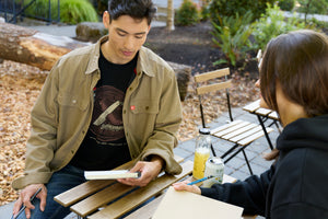 Translation missing: en.A man sitting at a table wearing a black Leatherman Blade &amp; Grain tee with a woman wearing a black hoodie, both equipped with a notebook and pencil