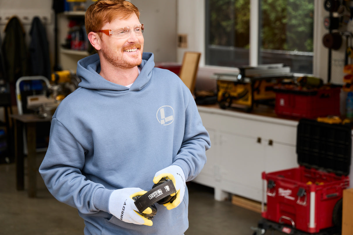 Translation missing: en.A person wearing a storm blue Leatherman Classic Hoodie smiling while holding headphones in a workshop