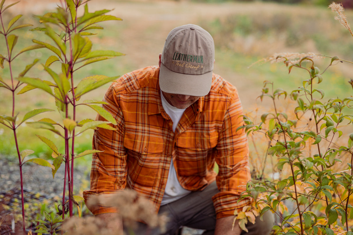 Translation missing: en.A person wearing a driftwood colored Leatherman Dad Hat tending to plants outdoors while dressed in an orange plaid shirt