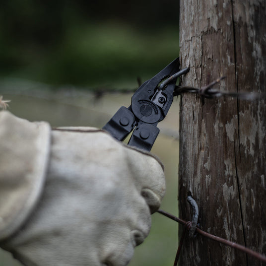 Translation missing: en.Gloved hand using a Leatherman Super Tool 300 multi-tool to tighten barbed wire on a wooden fence post, demonstrating rugged performance for agriculture and ranch work
