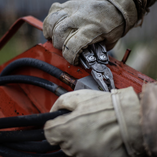 Translation missing: en.Gloved hands using a Leatherman Surge multi-tool to grip and adjust equipment on farm machinery, highlighting heavy-duty performance for agriculture tasks