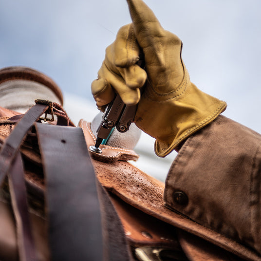 Translation missing: en.Person wearing yellow leather work gloves using a Leatherman Wave Plus multi-tool to tighten hardware on a leather saddle, demonstrating dependable tools for agriculture and ranch work