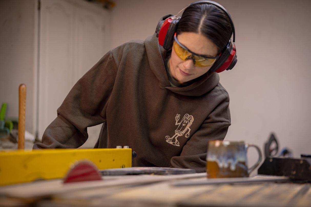 Translation missing: en.A person wearing a brown Leatherman Mr. Wave hoodie operating a table saw in a workshop, wearing safety glasses and earmuffs for protection