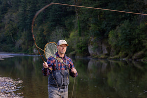 Translation missing: en.A person wearing a beige Leatherman PNW Trucker hat fly-fishing in a river surrounded by trees