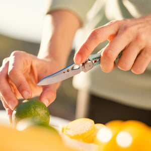 Translation missing: en.Person using an aqua Skeletool KB folding knife to slice a lime on a cutting surface outdoors