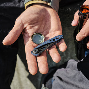 Translation missing: en.Hand holding a blue Skeletool KBX and bottle cap next to a person gripping an open glass bottle