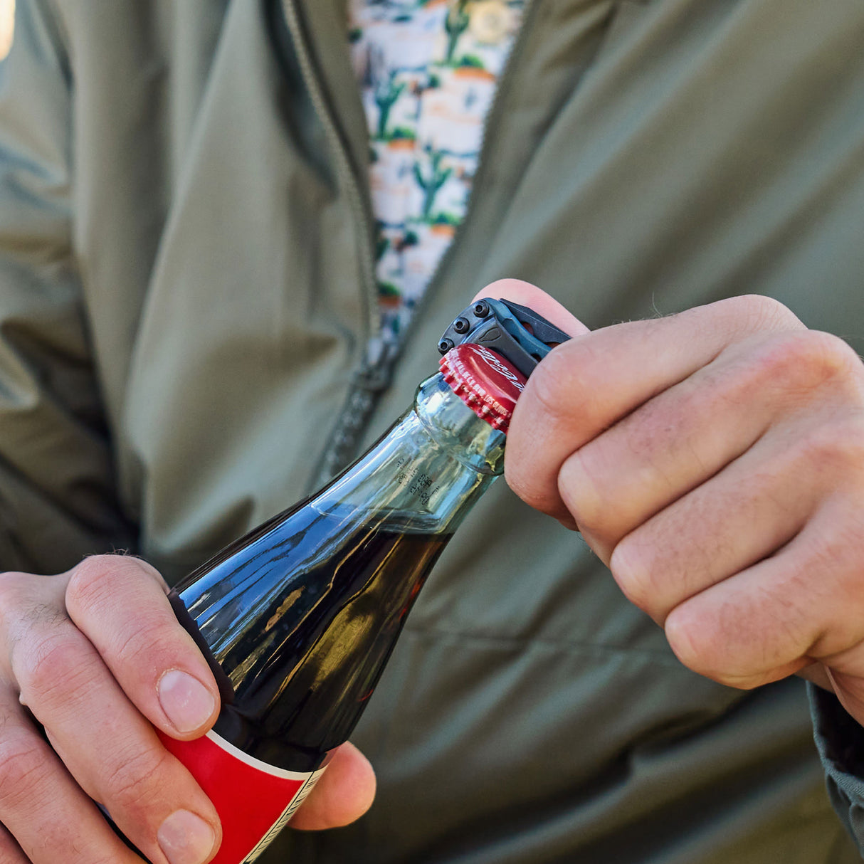 Translation missing: en.Hand using a Skeletool KBX to pry off the cap of a glass soda bottle