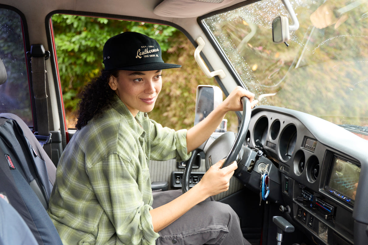 Translation missing: en.A woman wearing a black Leatherman Vintage 5-Panel Hat sitting in the driver’s seat of a vehicle, smiling while holding the steering wheel