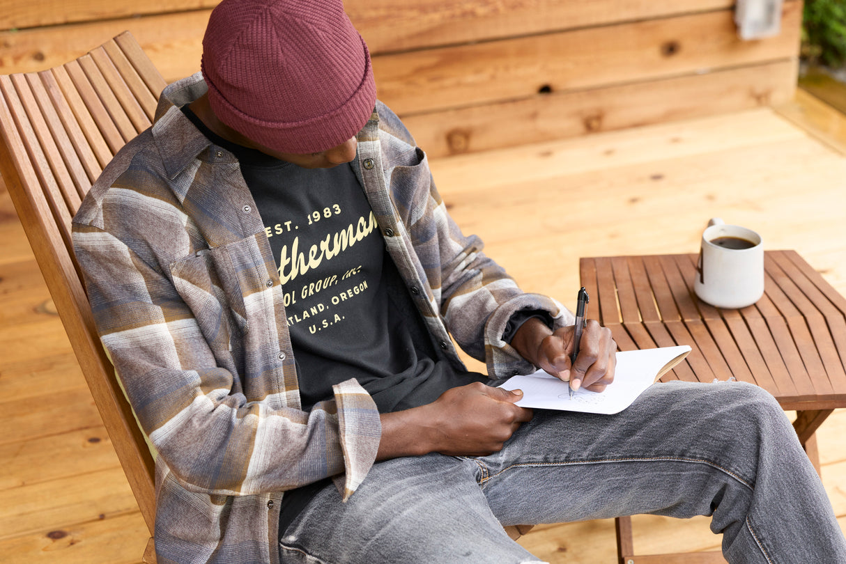 Translation missing: en.A person wearing a black Leatherman Vintage Tee sitting on a wooden chair outdoors, writing in a notebook with a mug of coffee on a nearby table