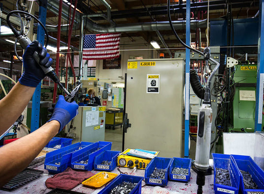 Translation missing: en.Leatherman tools being assembled on a manufacturing floor with workers, equipment, and an American flag in the background, highlighting USA manufacturing and craftsmanship