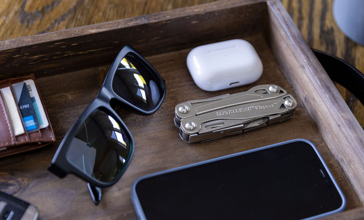 Translation missing: en.Stainless steel Leatherman Wingman® multi-tool beside sunglasses, smartphone, and AirPod case in a wooden tray
